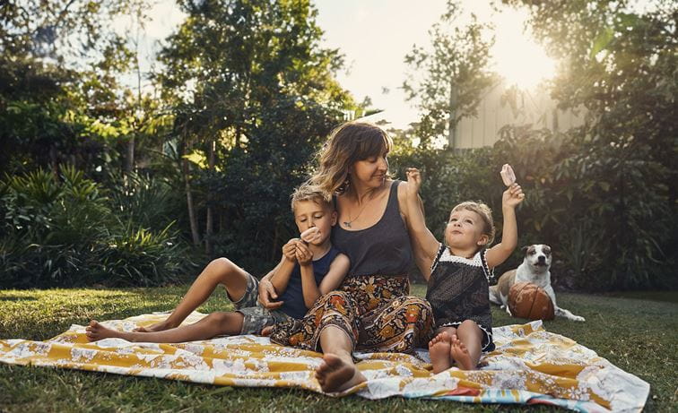 A woman shares a blanket with two children in a sunny backyward while they all eat ice-cream. A dog sits in the background.
