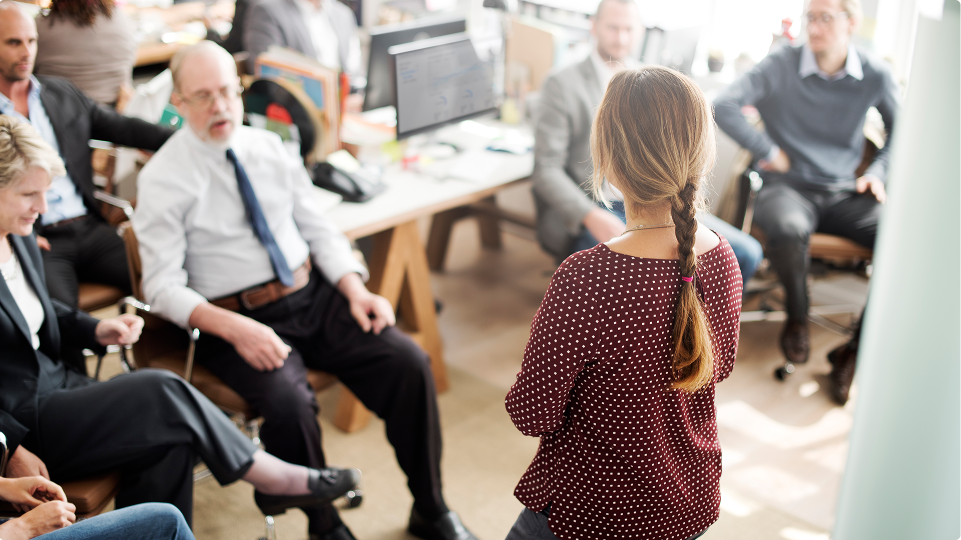 A woman stands at the front of a room of professional looking people. They are paying attention to her and she is explaining something to them.