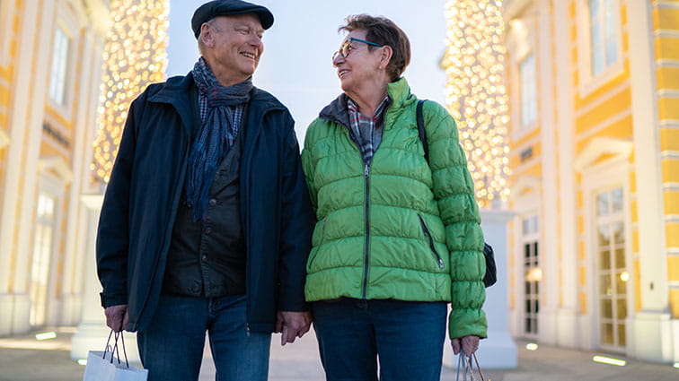 A man and woman are standing in a shopping district. Columns of lights are shining behind them and they are both holding shopping bags. They look at each other and smile like they are enjoying their shopping trip very much.